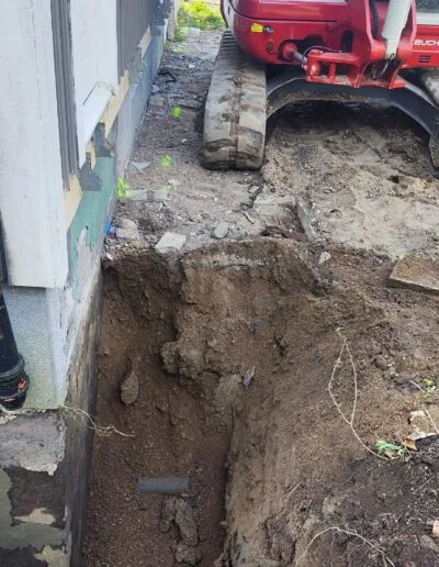 En bild på ett pågående dräneringsarbete där det syns en liten grävmaskin som används för att gräva upp runt husets grund.
